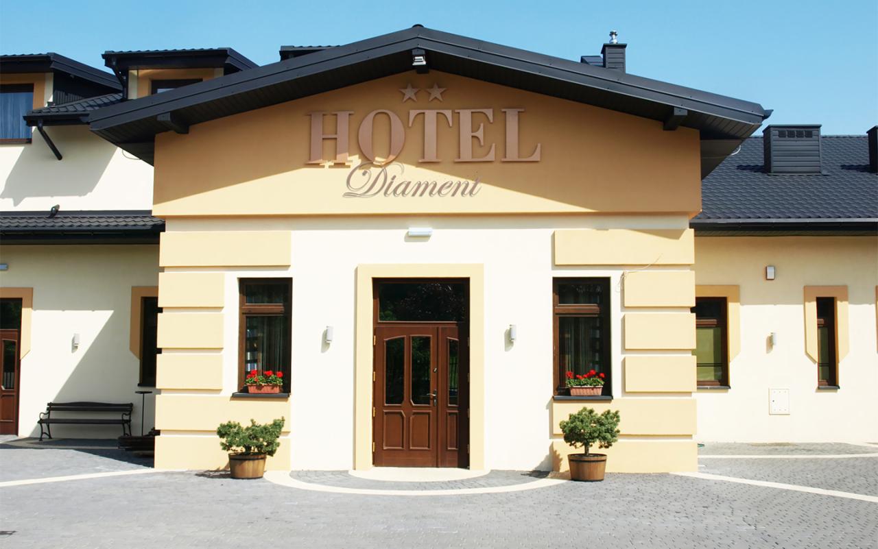 Elewacja budynku hotelu Diament z brązowymi oknami, drzwiami i napisem HOTEL nad wejściem, ozdobiona kwiatami w donicach i małymi drzewkami w drewnianych beczkach.