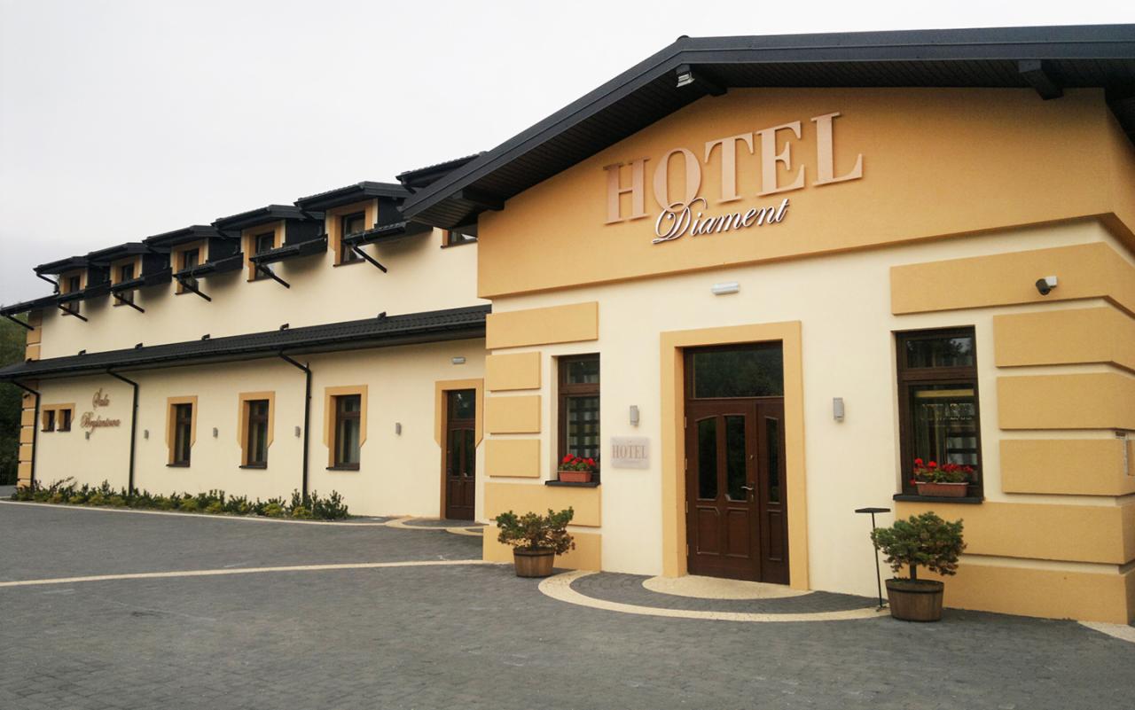 Elewacja budynku hotelu z napisem 'Hotel Diament' nad wejściem, widoczne okna z kwiatami i dekoracyjne krzewy w donicach.