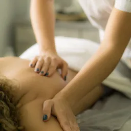 Terapeuta w białym uniformie wykonuje masaż pleców pacjentki z kręconymi włosami, widoczne dłonie z ciemnym lakierem do paznokci i ślady olejku.