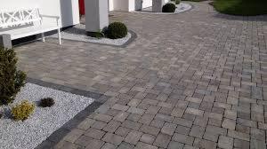 Ułożona kostka brukowa w odcieniach szarości, tworząca wzór na podjeździe, obrzeża z białego grysu i niskie krzewy ozdobne wzdłuż ścieżki.