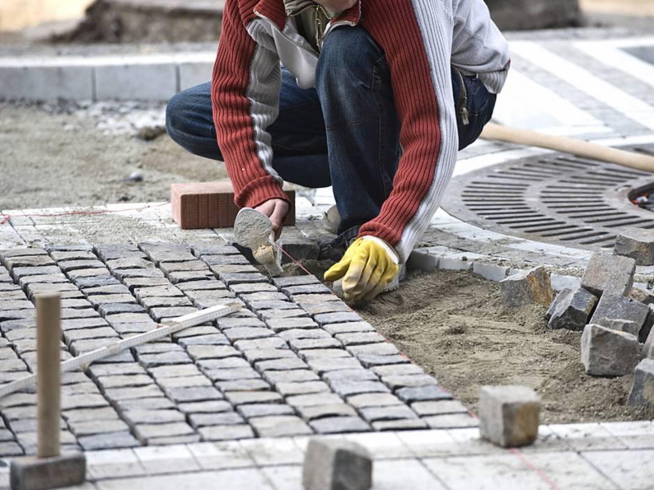 Pracownik w żółtych rękawicach układa kostkę brukową, wyrównując ją za pomocą kielni i piasku, z widoczną czerwoną linką wyznaczającą linię, cegłą oraz fragmentem studzienki kanalizacyjnej w tle.