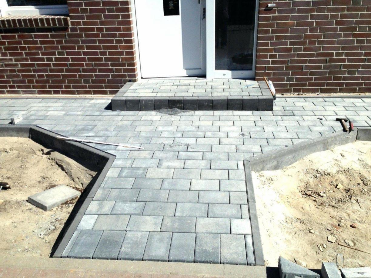 Układanie szarej kostki brukowej przed wejściem do budynku z cegły, widoczne narzędzia i piasek, etap prac wykończeniowych.