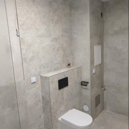 NOVEX Sławomir Nowak - Nowoczesna łazienka z podwieszaną toaletą, wykończona płytkami w odcieniach szarości z delikatnym wzorem, widoczny fragment kabiny prysznicowej z chromowanym wieszakiem.