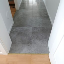 NOVEX Sławomir Nowak - Długi korytarz wyłożony dużymi, szarymi płytkami imitującymi beton, z widocznym przejściem do pomieszczenia z jasnymi panelami podłogowymi oddzielonymi złotą listwą progową.