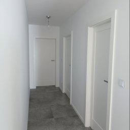 NOVEX Sławomir Nowak - Korytarz w jasnych barwach z szarą podłogą z płytek imitujących kamień, trzema białymi drzwiami i minimalistyczną lampą sufitową.