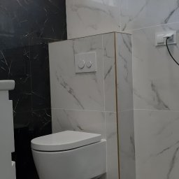 NOVEX Sławomir Nowak - Nowoczesna toaleta podwieszana z białymi płytkami imitującymi marmur, czarne płytki z marmurowym wzorem po lewej stronie, szare płytki podłogowe i złote akcenty przy krawędziach płytek.