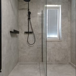 NOVEX Sławomir Nowak - Nowoczesna kabina prysznicowa typu walk-in z deszczownicą w kolorze czarnym, szklaną ścianką i odpływem liniowym, wykończona płytkami w odcieniach szarości, oświetlona przez okno z żaluzjami.