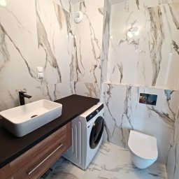 NOVEX Sławomir Nowak - Nowoczesna łazienka w Dobroszycach z marmurowymi płytkami, umywalką nablatową z czarną armaturą, drewnianą szafką pod umywalką, pralką i podwieszaną toaletą.