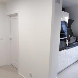 NOVEX Sławomir Nowak - Jasne wnętrze po remoncie: widok na białą ścianę z gniazdkiem, drzwi wewnętrzne i fragment zabudowy kuchennej z piekarnikiem i szafkami.