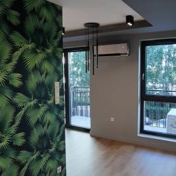 NOVEX Sławomir Nowak - Nowoczesne wnętrze po remoncie: ściana z tapetą w liście, drewniana podłoga, czarne lampy sufitowe i klimatyzacja.