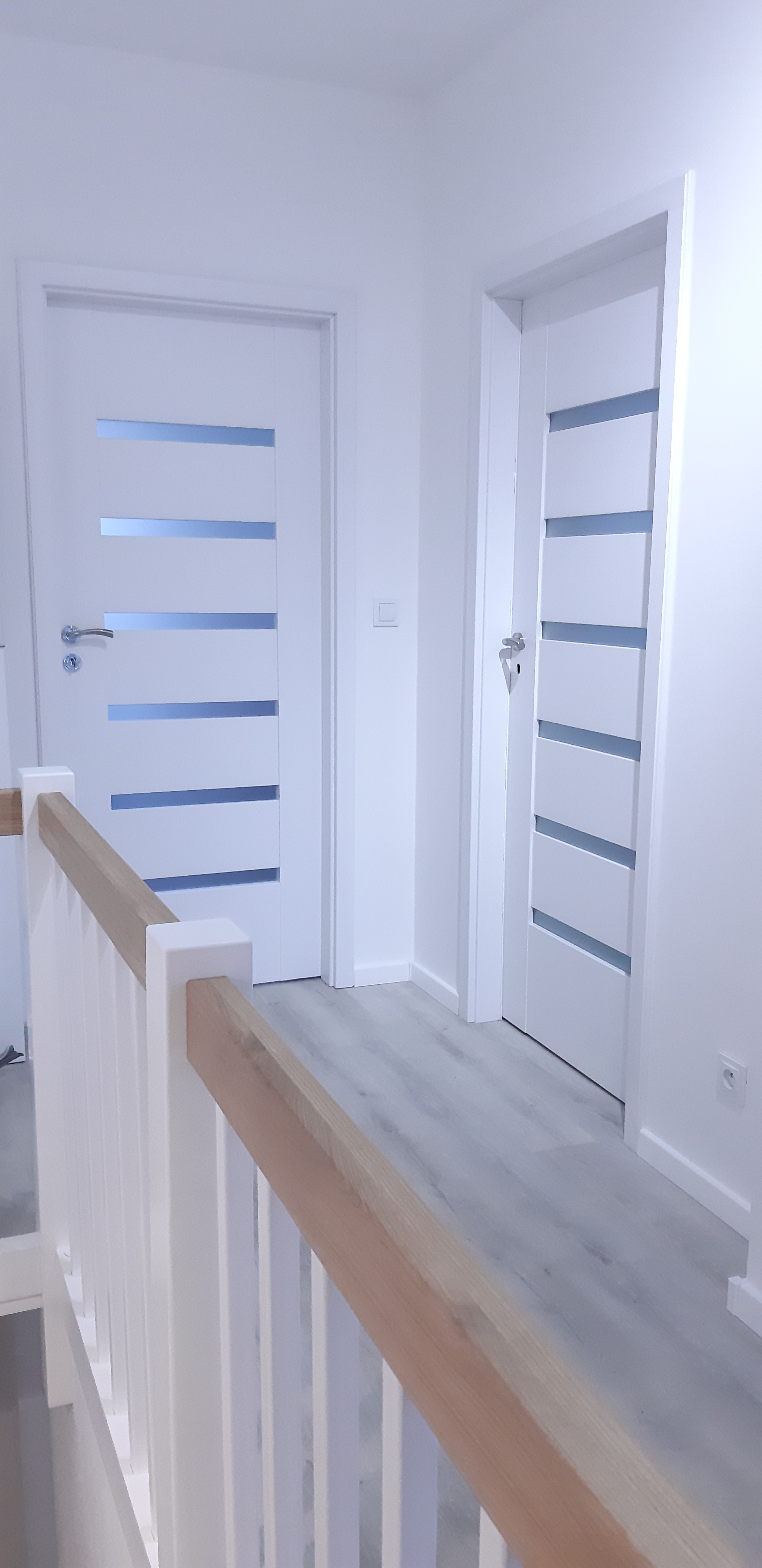 Nowoczesne, białe drzwi wewnętrzne z poziomymi przeszkleniami, widok z góry schodów z drewnianą balustradą, szara podłoga imitująca drewno.