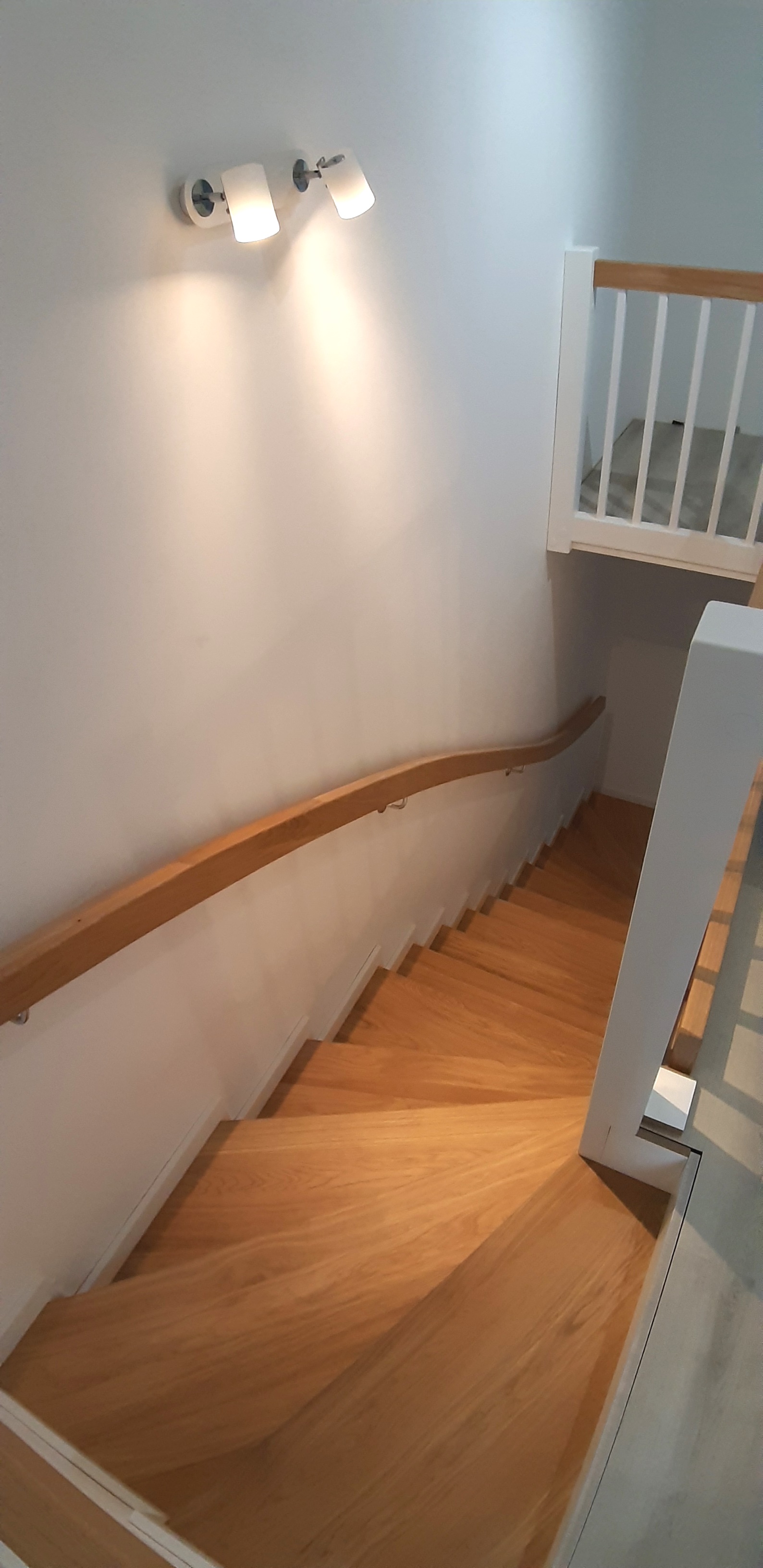 Drewniane schody z jasną balustradą w domu w Dobroszycach, oświetlone lampami ściennymi, widok z góry na zakręcające stopnie.