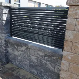 Nowoczesne ogrodzenie z poziomych, czarnych profili zamontowane między słupkami z szarego i beżowego kamienia łupanego, z widocznym piaskiem na kostce brukowej.