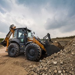 REY-KOP Miłosz Baleja - Żółto-czarna koparko-ładowarka CASE nabiera ziemię z hałdy kamieni na placu budowy, widoczne ślady opon na piaszczystym podłożu, pochmurne niebo w tle.