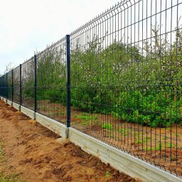 REY-KOP Miłosz Baleja - Długi, czarny panelowy płot z podmurówką z imitacji kamienia, ciągnący się wzdłuż granicy działki z widocznymi drzewami owocowymi w tle, świeżo obsypany ziemią.