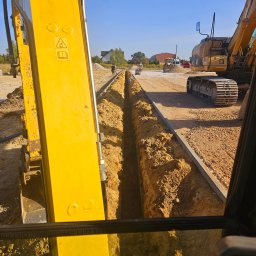 REY-KOP Miłosz Baleja - Wykopany wąski rów w ziemi, widok z kabiny żółtej koparki, w tle pracownicy i inne maszyny budowlane, ułożone krawężniki po bokach wykopu.