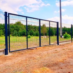 REY-KOP Miłosz Baleja - Nowoczesne, ciemne ogrodzenie panelowe z bramą wjazdową, osadzone na betonowej podmurówce, z widokiem na zielone drzewa w tle i piaszczystą nawierzchnią na pierwszym planie.