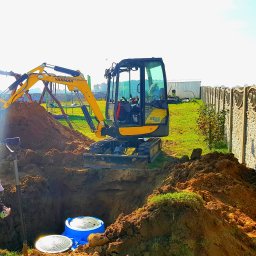 REY-KOP Miłosz Baleja - Żółta mini koparka Yanmar podczas wykopu pod zbiornik z niebieskim wiekiem, widoczna łopata, ziemia i trawa, w tle ogrodzenie betonowe i budynek gospodarczy.