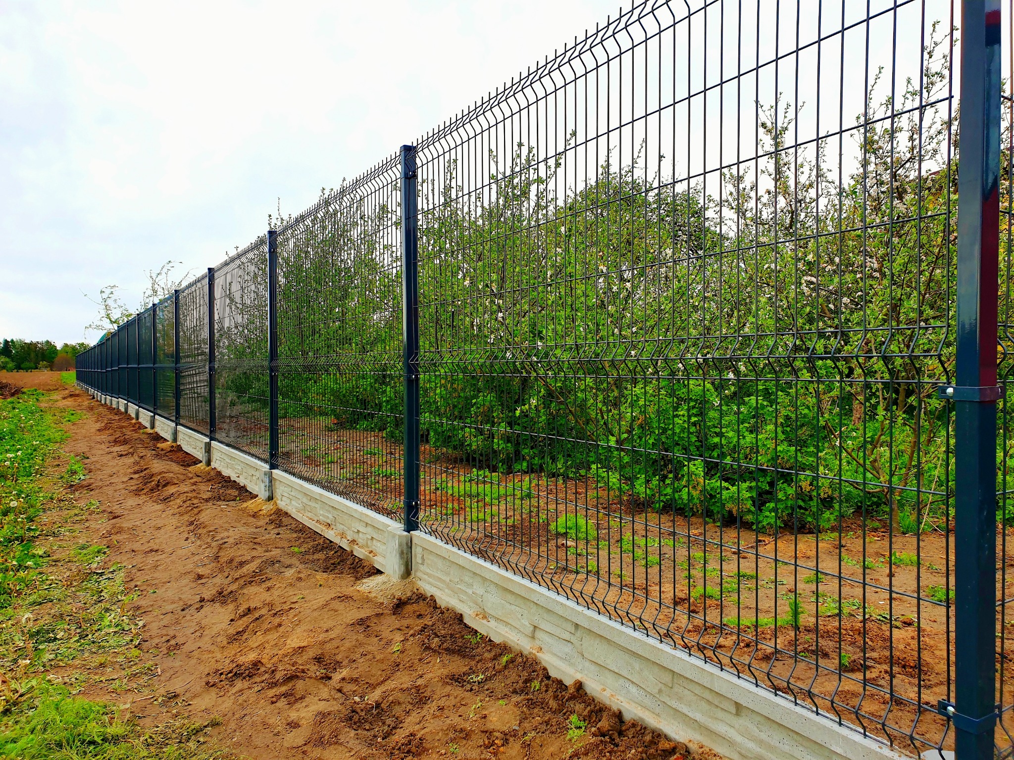 Długi, czarny panelowy płot z podmurówką z imitacji kamienia, ciągnący się wzdłuż granicy działki z widocznymi drzewami owocowymi w tle, świeżo obsypany ziemią.