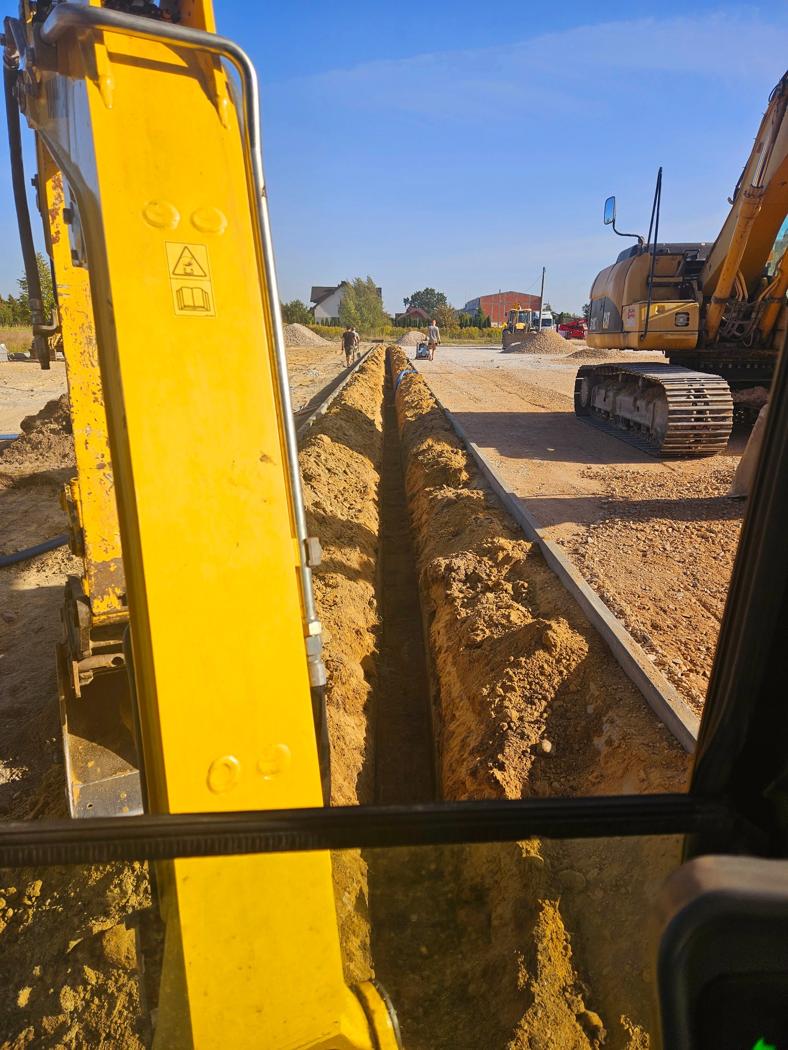 Wykopany wąski rów w ziemi, widok z kabiny żółtej koparki, w tle pracownicy i inne maszyny budowlane, ułożone krawężniki po bokach wykopu.