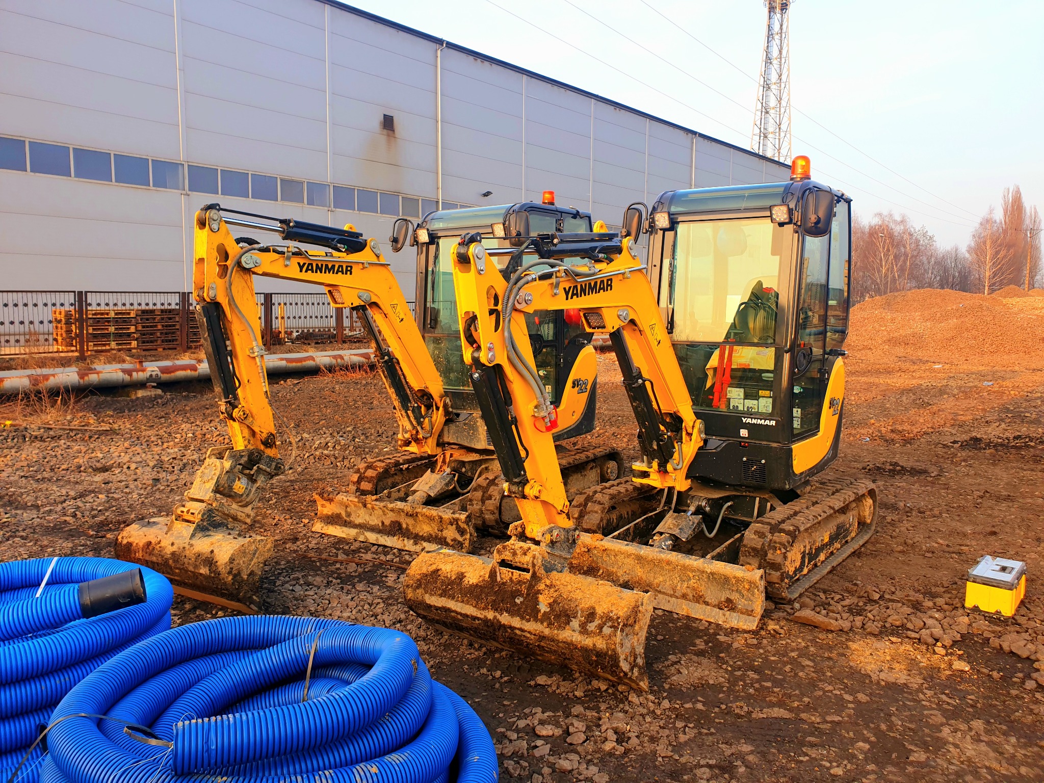 Dwa żółte mini-koparki Yanmar stojące na placu budowy z stertą niebieskich rur w tle, widoczne budynki przemysłowe i hałda ziemi.