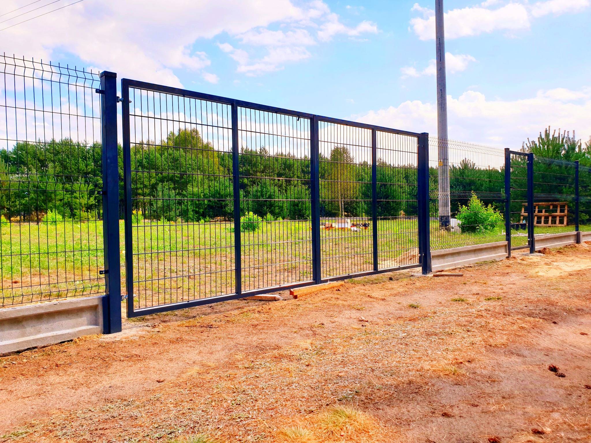 Nowoczesne, ciemne ogrodzenie panelowe z bramą wjazdową, osadzone na betonowej podmurówce, z widokiem na zielone drzewa w tle i piaszczystą nawierzchnią na pierwszym planie.