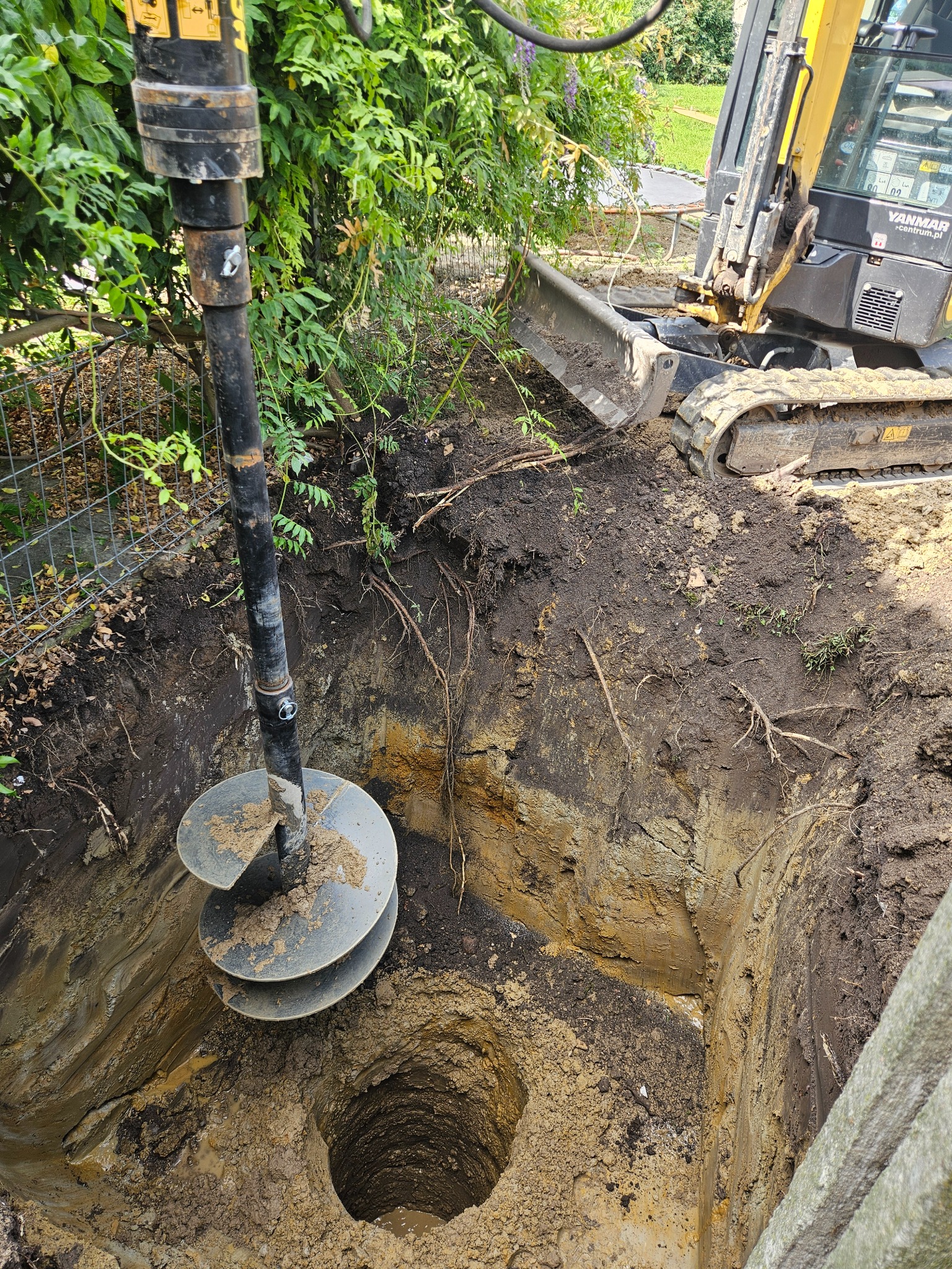 Żółty minikoparka Yanmar z wiertnicą hydrauliczną wierci głęboki otwór w ziemi o warstwowej strukturze gleby, widoczne korzenie i resztki roślinności.