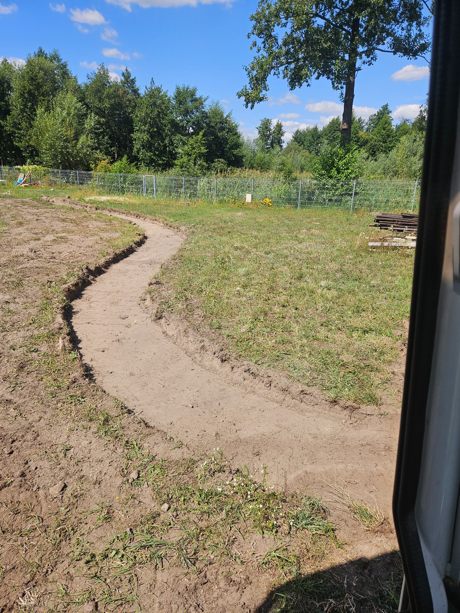 Wykopany, wijący się rów w trawiastym terenie, widok zza krawędzi pojazdu, drzewa i ogrodzenie w tle.
