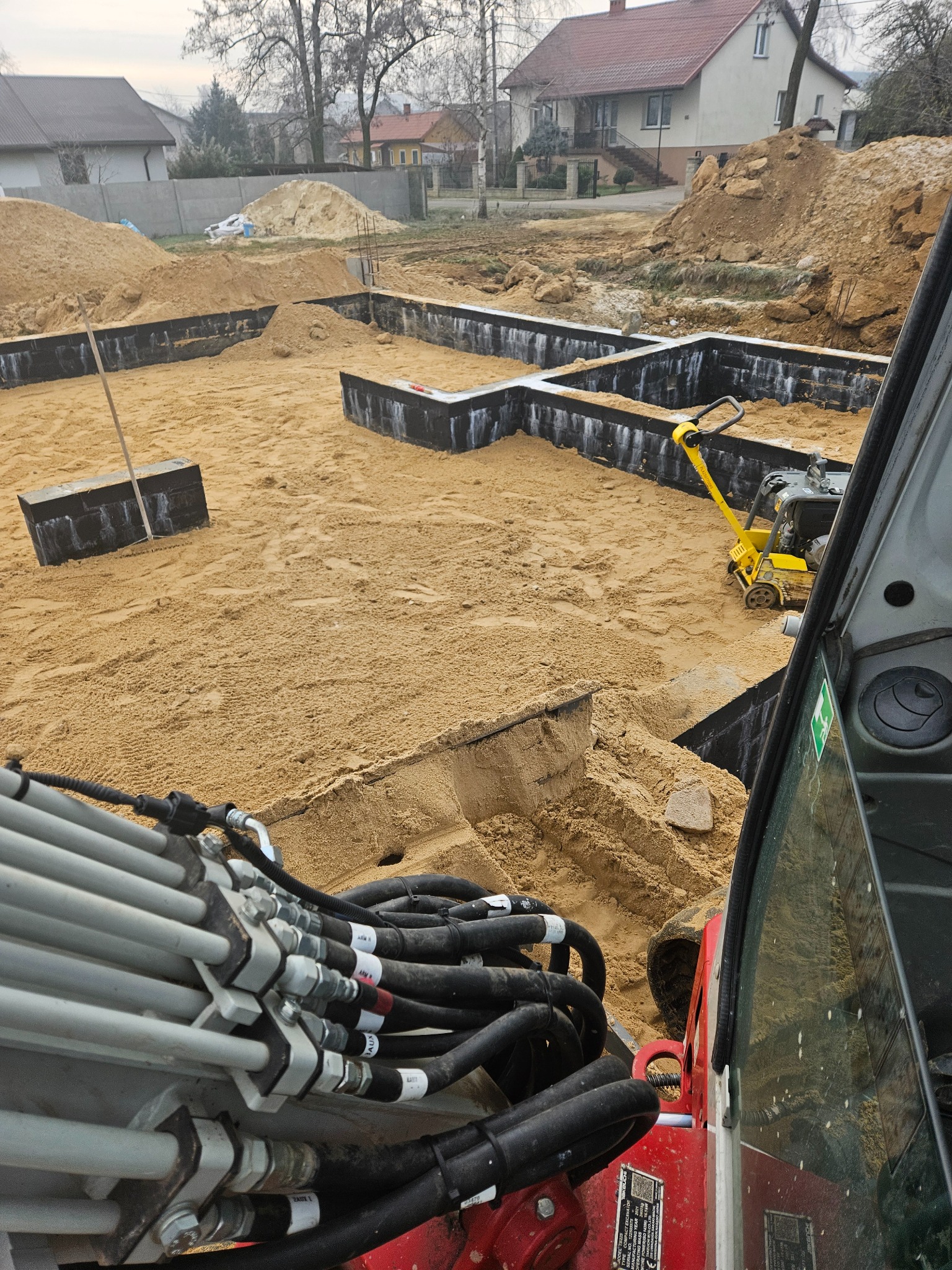 Przygotowanie terenu pod fundamenty domu jednorodzinnego: wykop z szalunkiem, piasek zasypowy, ubijarka wibracyjna i widoczne przewody hydrauliczne koparki.
