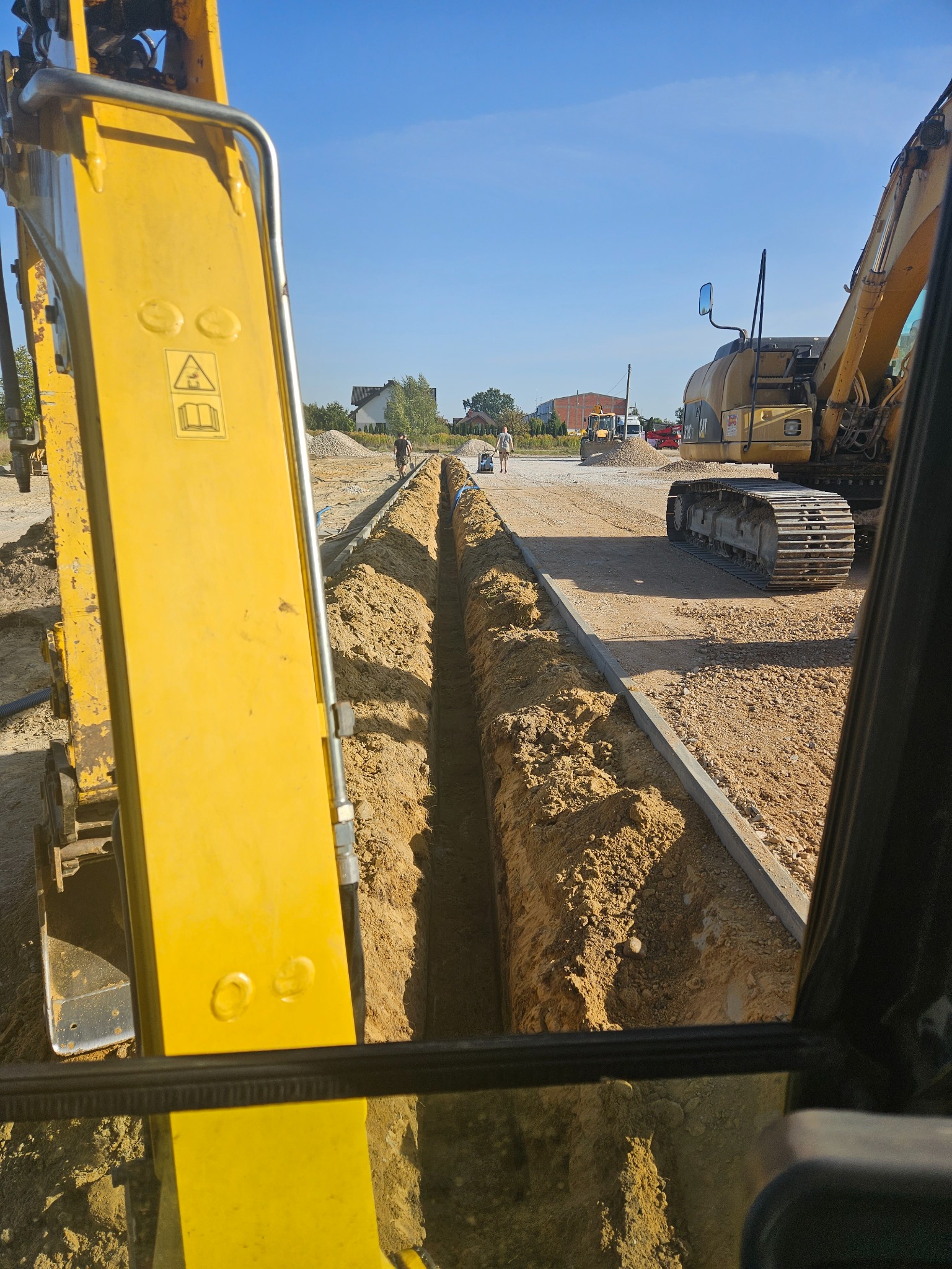 Głęboki wykop liniowy z ułożonymi rurami, widok z kabiny koparki, w tle pracownicy i inny sprzęt budowlany.