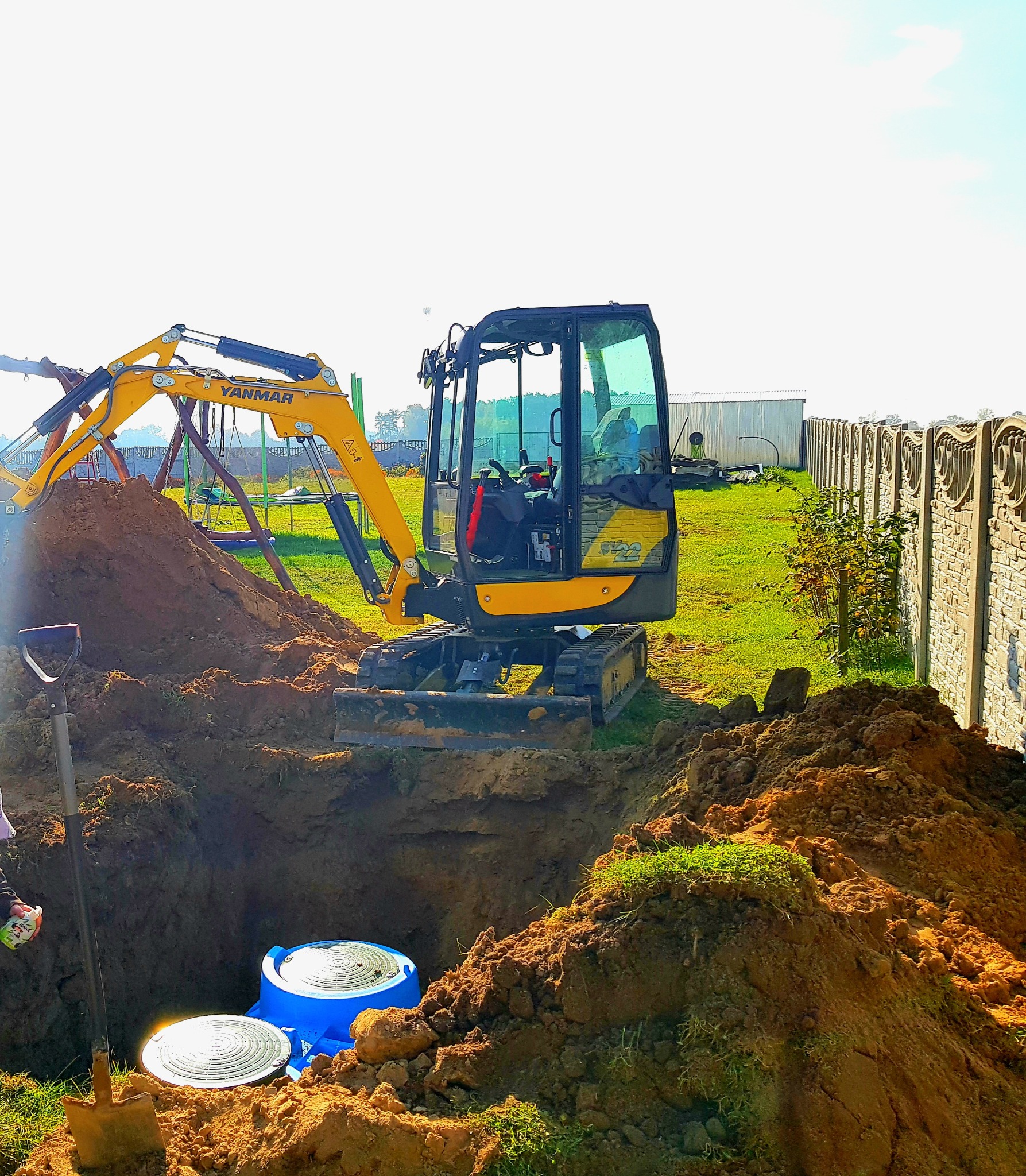 Żółta mini koparka Yanmar podczas wykopu pod zbiornik z niebieskim wiekiem, widoczna łopata, ziemia i trawa, w tle ogrodzenie betonowe i budynek gospodarczy.