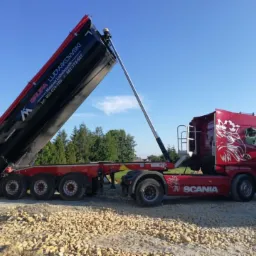 Czerwona ciężarówka Scania z otwartą naczepą wywrotką, wysypująca ładunek na żwirowy teren, widoczne logo firmy transportowej na naczepie.