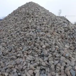 Duża pryzma szarego kruszywa o nieregularnych kształtach, widoczna na tle pochmurnego nieba i ogrodzenia przemysłowego.