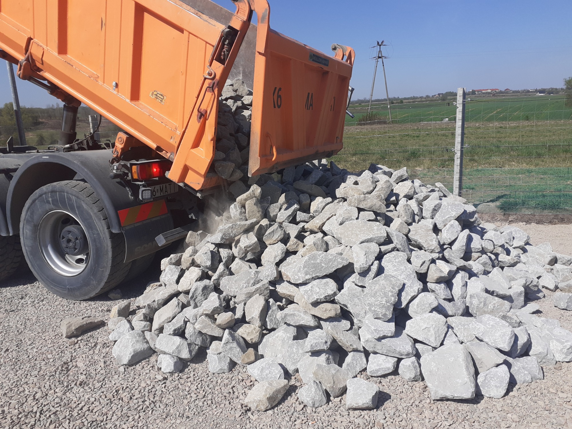 Pomarańczowa wywrotka wysypuje szare kamienie na szutrowe podłoże, widoczne pole w tle.