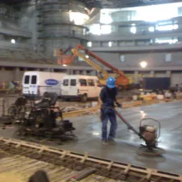 Pracownik w niebieskim kasku wygładza świeżo wylaną posadzkę za pomocą zacieraczki mechanicznej na budowie hali, w tle zaparkowane busy i podnośniki.