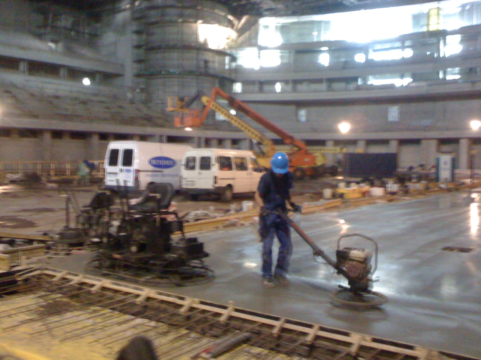 Pracownik w niebieskim kasku wygładza świeżo wylaną posadzkę za pomocą zacieraczki mechanicznej na budowie hali, w tle zaparkowane busy i podnośniki.