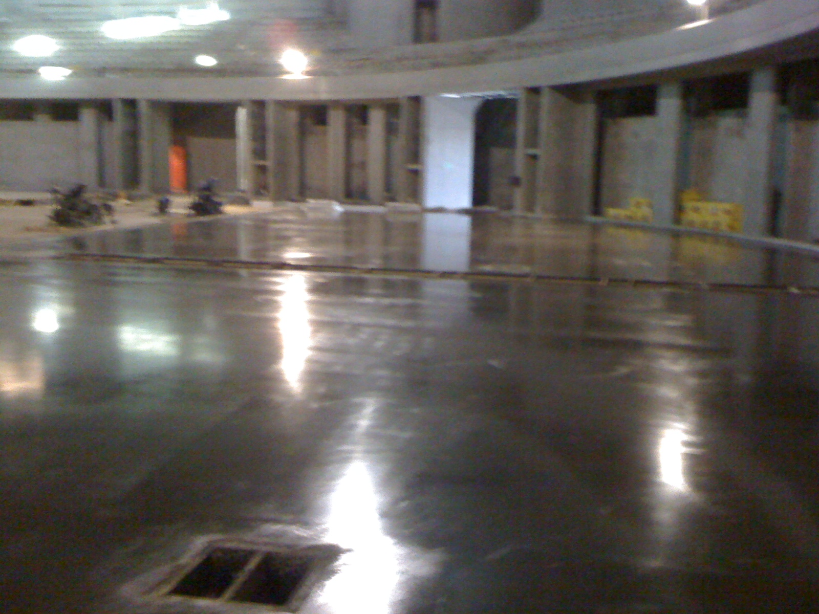 Błyszcząca, szara powierzchnia w hali przemysłowej, z widocznymi refleksami świetlnymi, częściowo wykończona, z pracownikami w tle i otworami w podłodze.