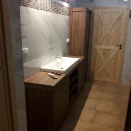 Nowoczesna łazienka z drewnianą zabudową umywalki, białym blatem i umywalką, płytkami imitującymi marmur na ścianach, mozaiką nad umywalką i drewnianymi drzwiami w stylu rustykalnym.