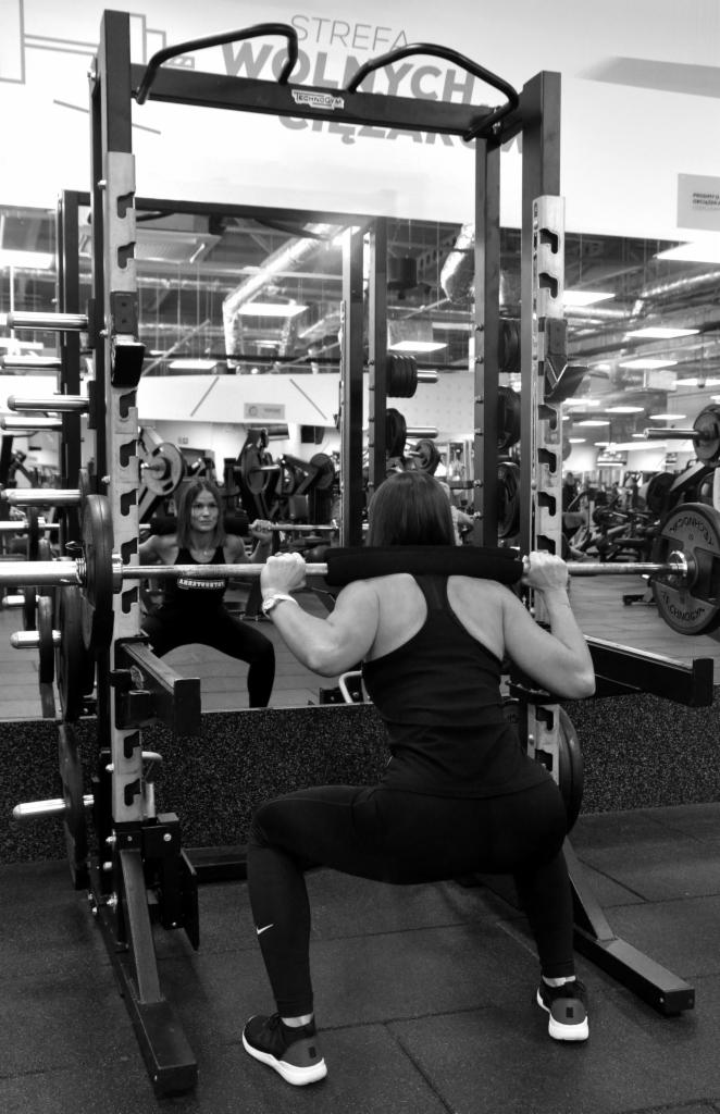 Kobieta wykonuje przysiad ze sztangą na suwnicy Smitha w siłowni, widoczne odbicie w lustrze, czarno-białe zdjęcie.