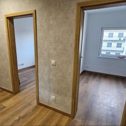 Dzorki - Wnętrze mieszkania po remoncie: drewnopodobna podłoga, ściany w odcieniach szarości, dwoje drzwi z drewnianymi ościeżnicami, widok na okno i budynek z balkonami.