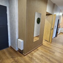 Dzorki - Nowoczesny przedpokój po remoncie: drewniane panele ścienne, lustro z roślinnym akcentem, ciemne drzwi wejściowe i widok na jasną kuchnię z drewnianym blatem.