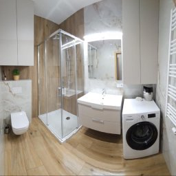 Dzorki - Nowoczesna łazienka po remoncie: kabina prysznicowa, umywalka z szafką, toaleta, pralka i grzejnik drabinkowy. Płytki imitujące drewno i marmur. Całość w jasnych, stonowanych barwach.