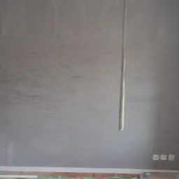 Ściana w szarym odcieniu z efektem dekoracyjnym, widoczna drabina malarska, zabezpieczona podłoga i wystające kable elektryczne.