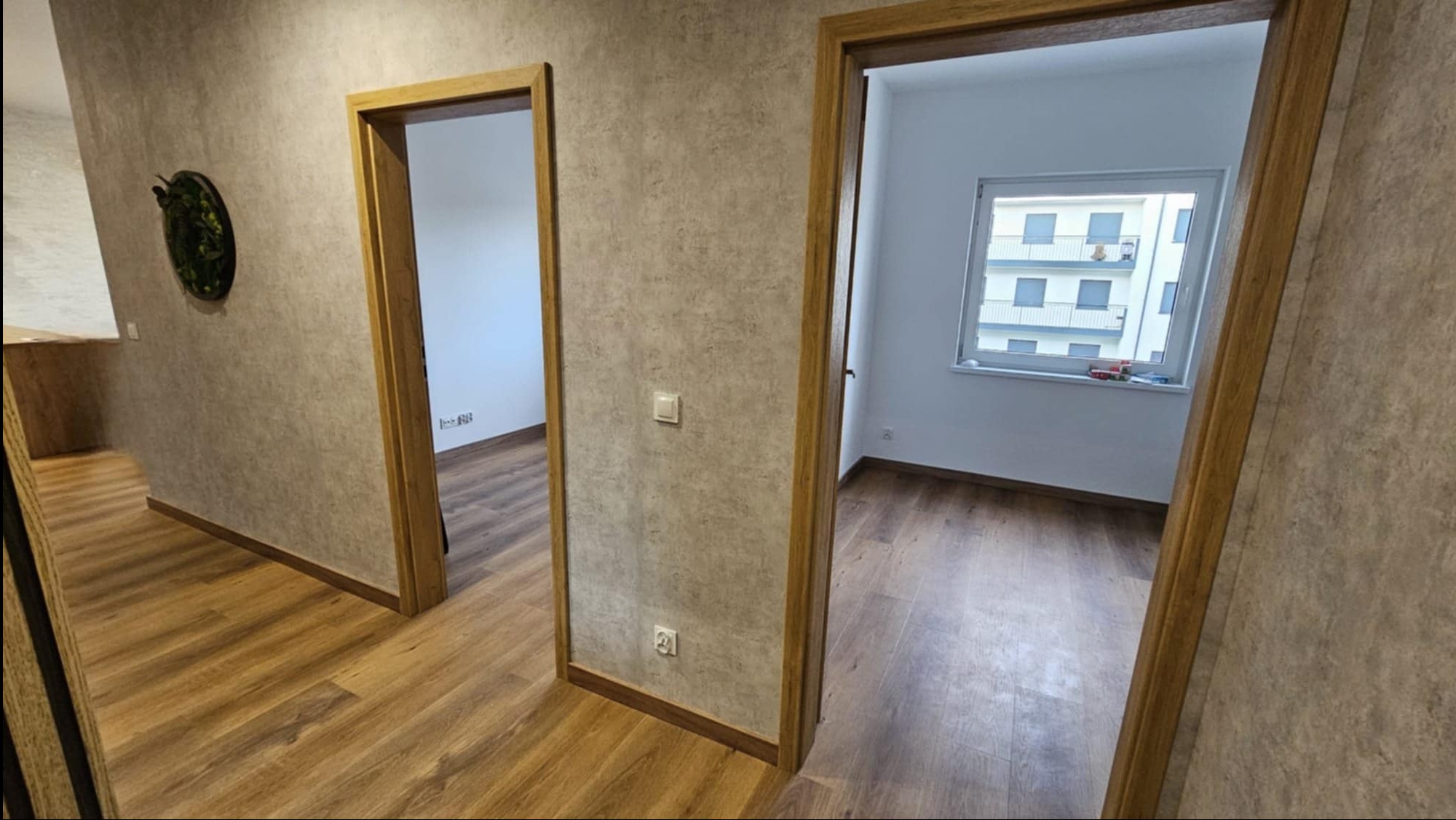Wnętrze mieszkania po remoncie: drewnopodobna podłoga, ściany w odcieniach szarości, dwoje drzwi z drewnianymi ościeżnicami, widok na okno i budynek z balkonami.