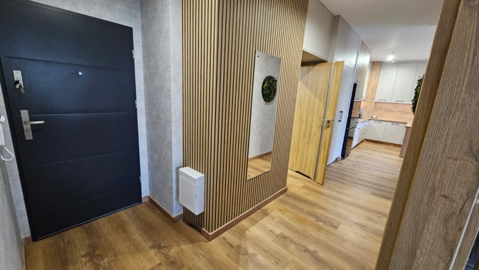 Nowoczesny przedpokój po remoncie: drewniane panele ścienne, lustro z roślinnym akcentem, ciemne drzwi wejściowe i widok na jasną kuchnię z drewnianym blatem.