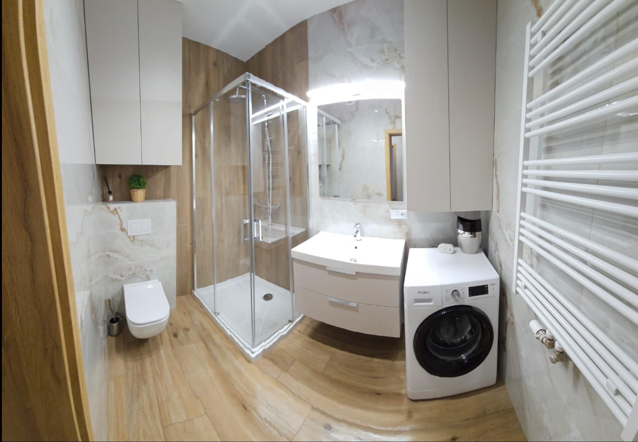 Nowoczesna łazienka po remoncie: kabina prysznicowa, umywalka z szafką, toaleta, pralka i grzejnik drabinkowy. Płytki imitujące drewno i marmur. Całość w jasnych, stonowanych barwach.