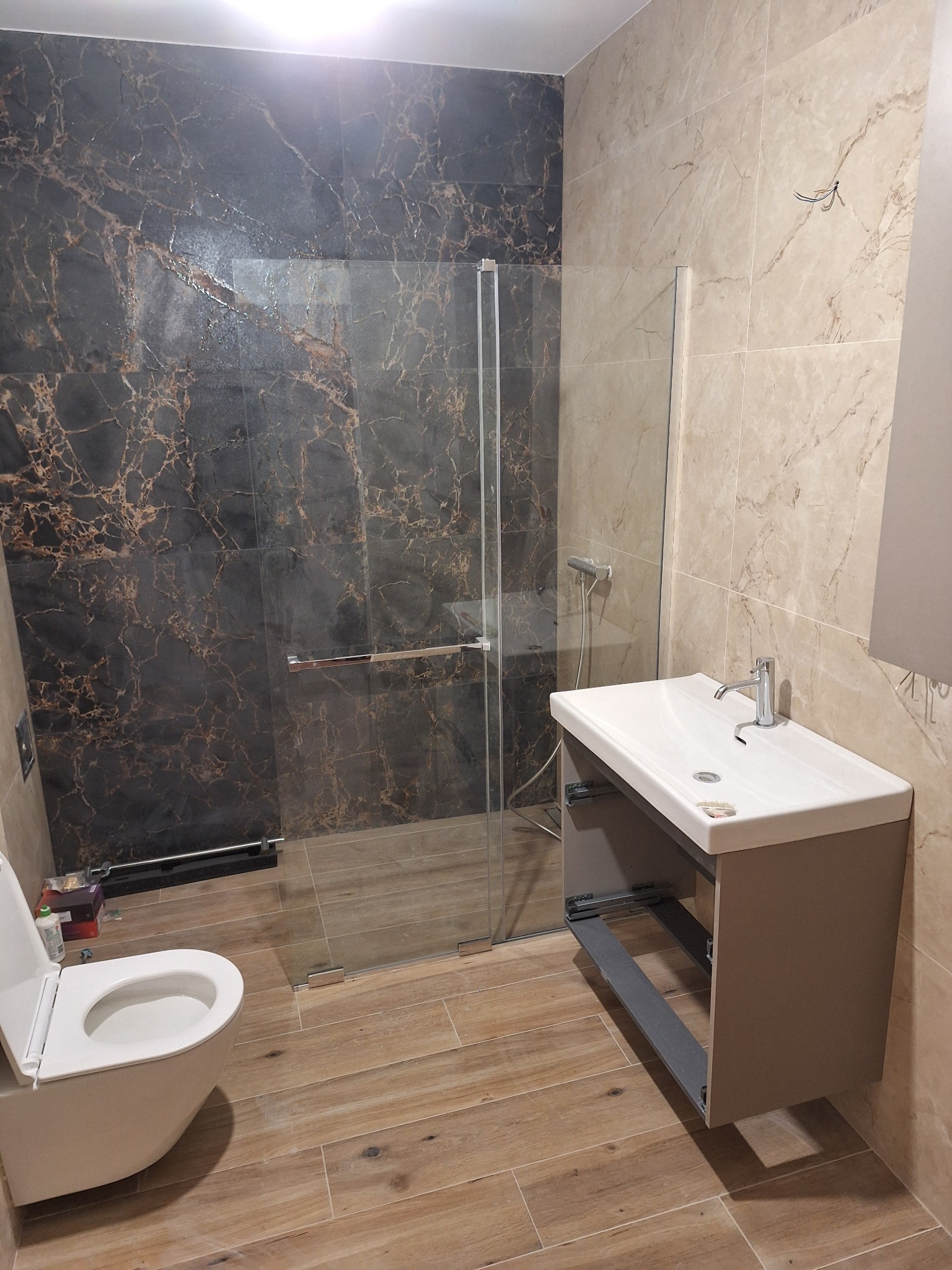 Wykończona łazienka w Nowym Tomyślu: kabina prysznicowa ze szklanymi drzwiami, umywalka z szafką i toaleta. Na ścianach płytki imitujące marmur.