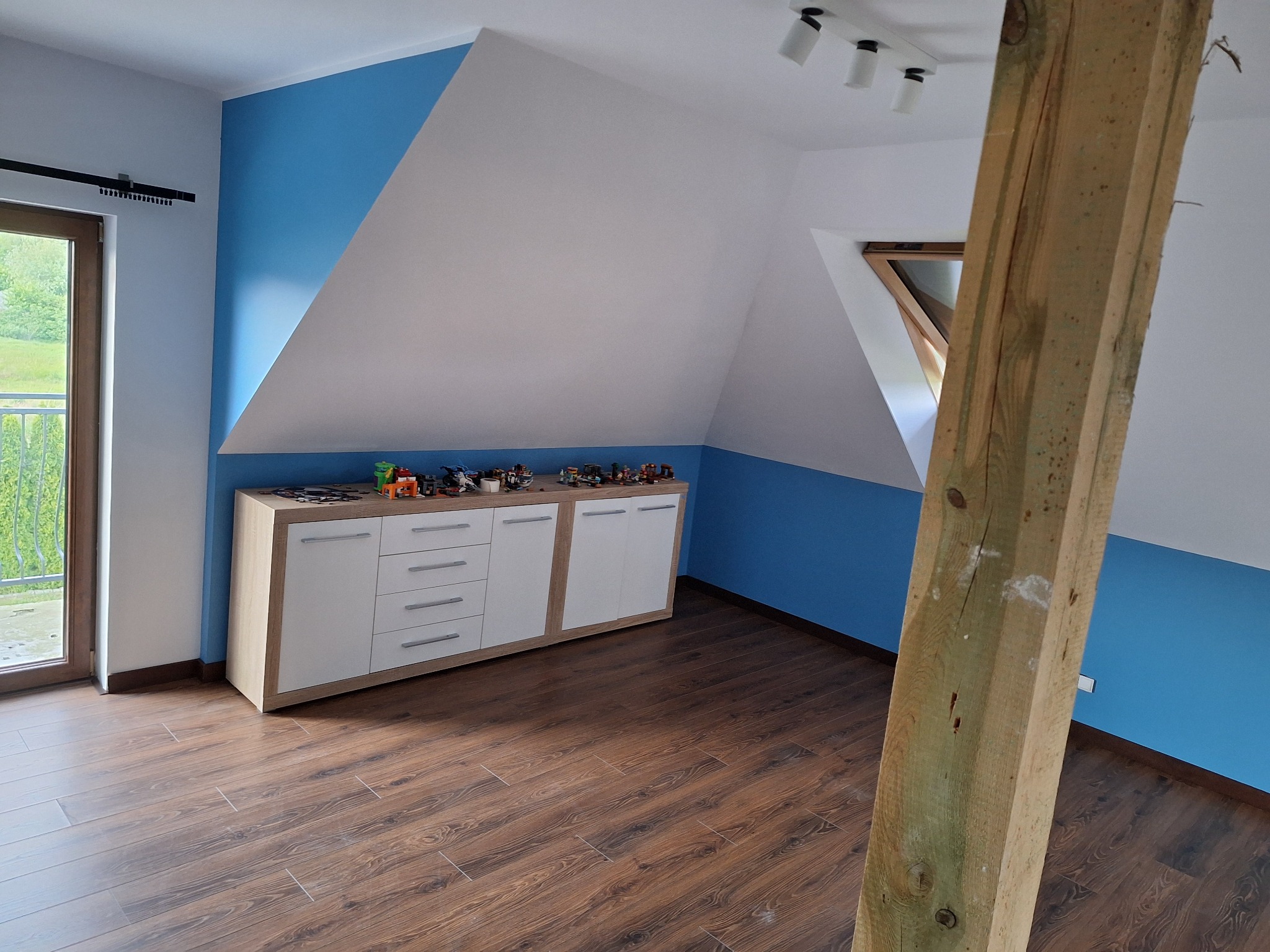 Pokój na poddaszu po remoncie: biało-niebieskie ściany, drewniana podłoga, biała komoda z zabawkami, okno dachowe i drewniany słup konstrukcyjny.