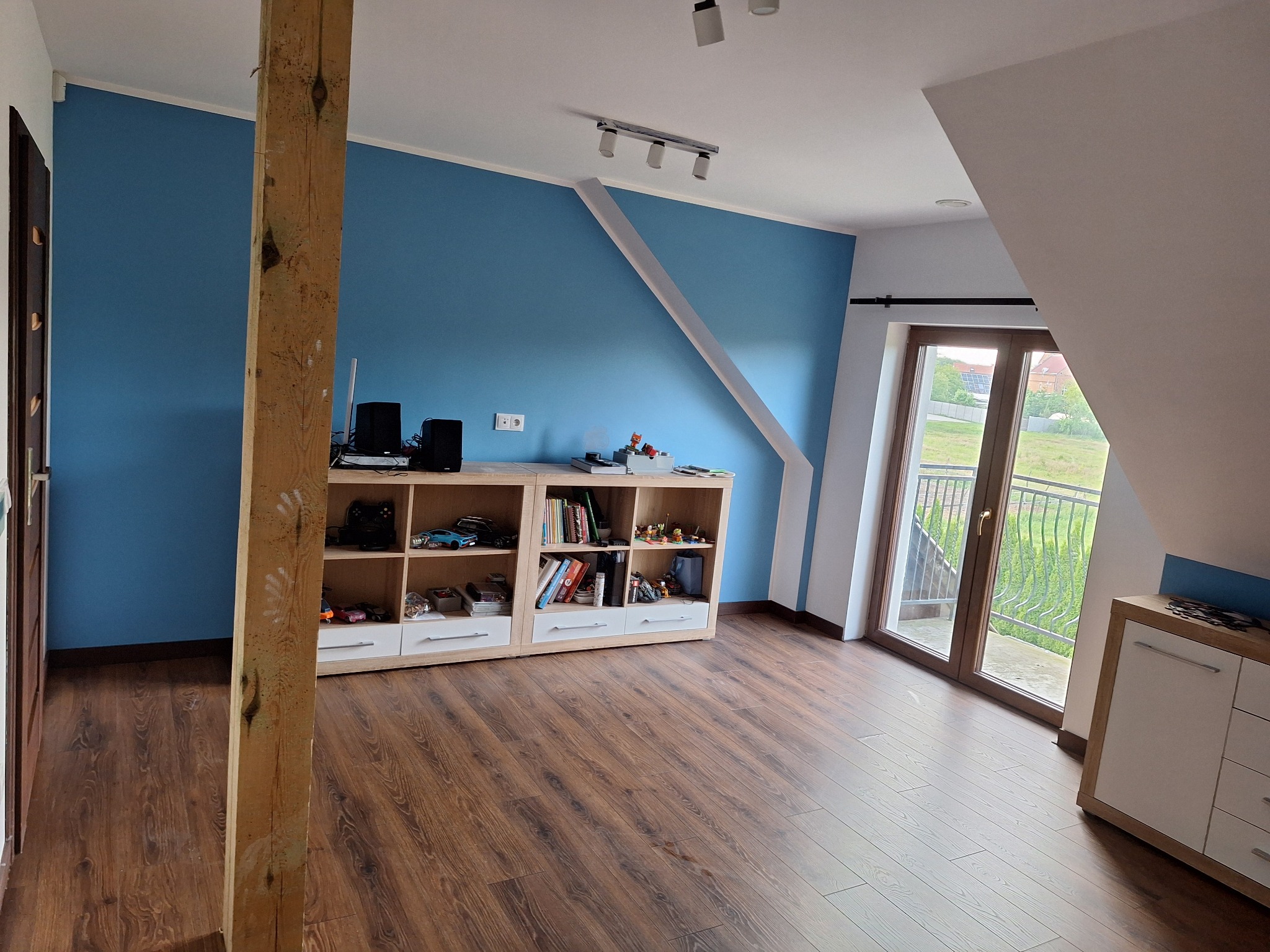 Błękitna ściana w pokoju na poddaszu z drewnianą podłogą i regałem. Widoczny fragment drewnianej kolumny i okno balkonowe z widokiem na zieleń.