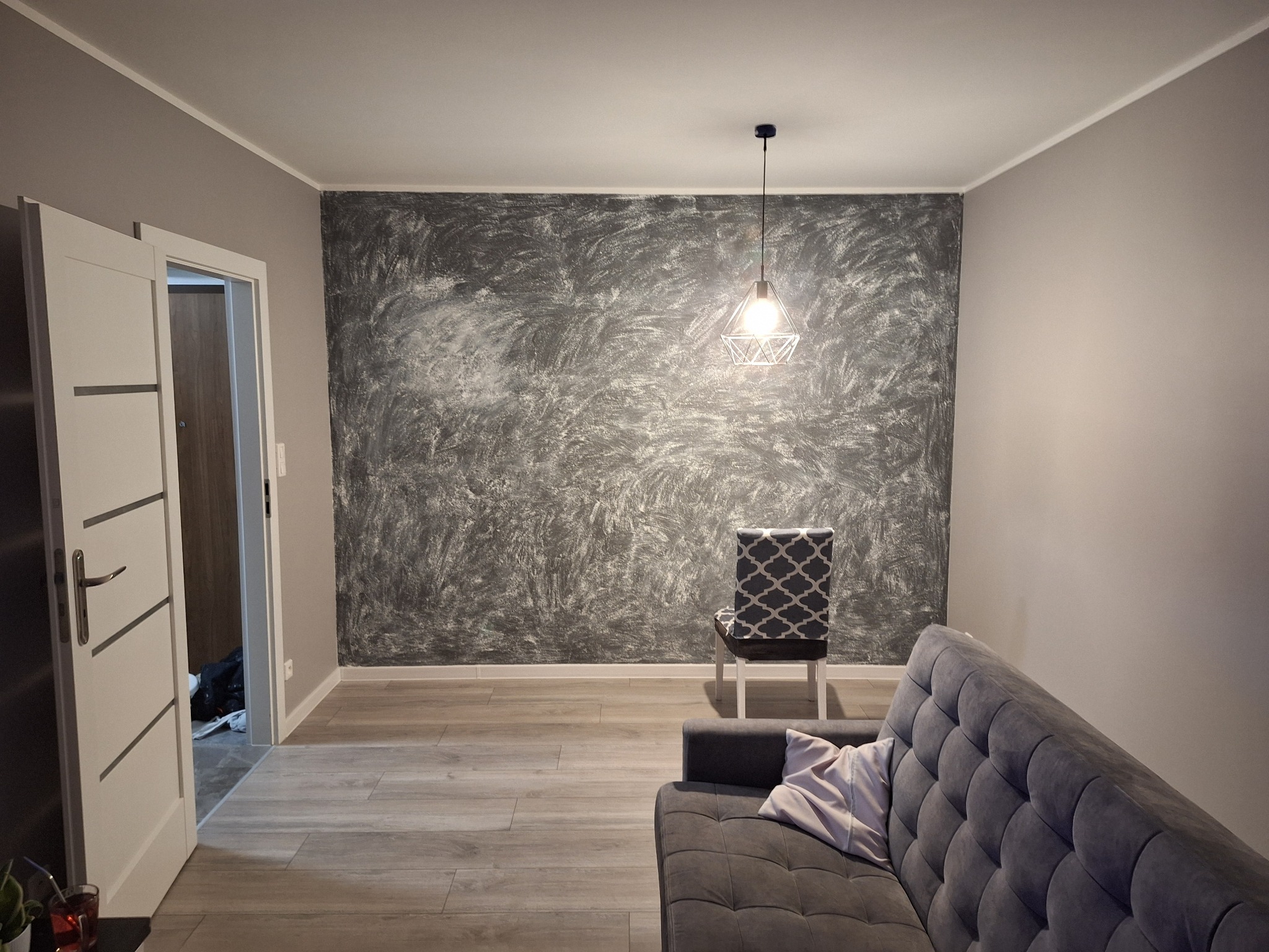 Stylowe wnętrze pokoju po odświeżeniu: szara ściana z unikalną fakturą, nowoczesna lampa i wygodna sofa. Widok na otwarte drzwi do innego pomieszczenia.